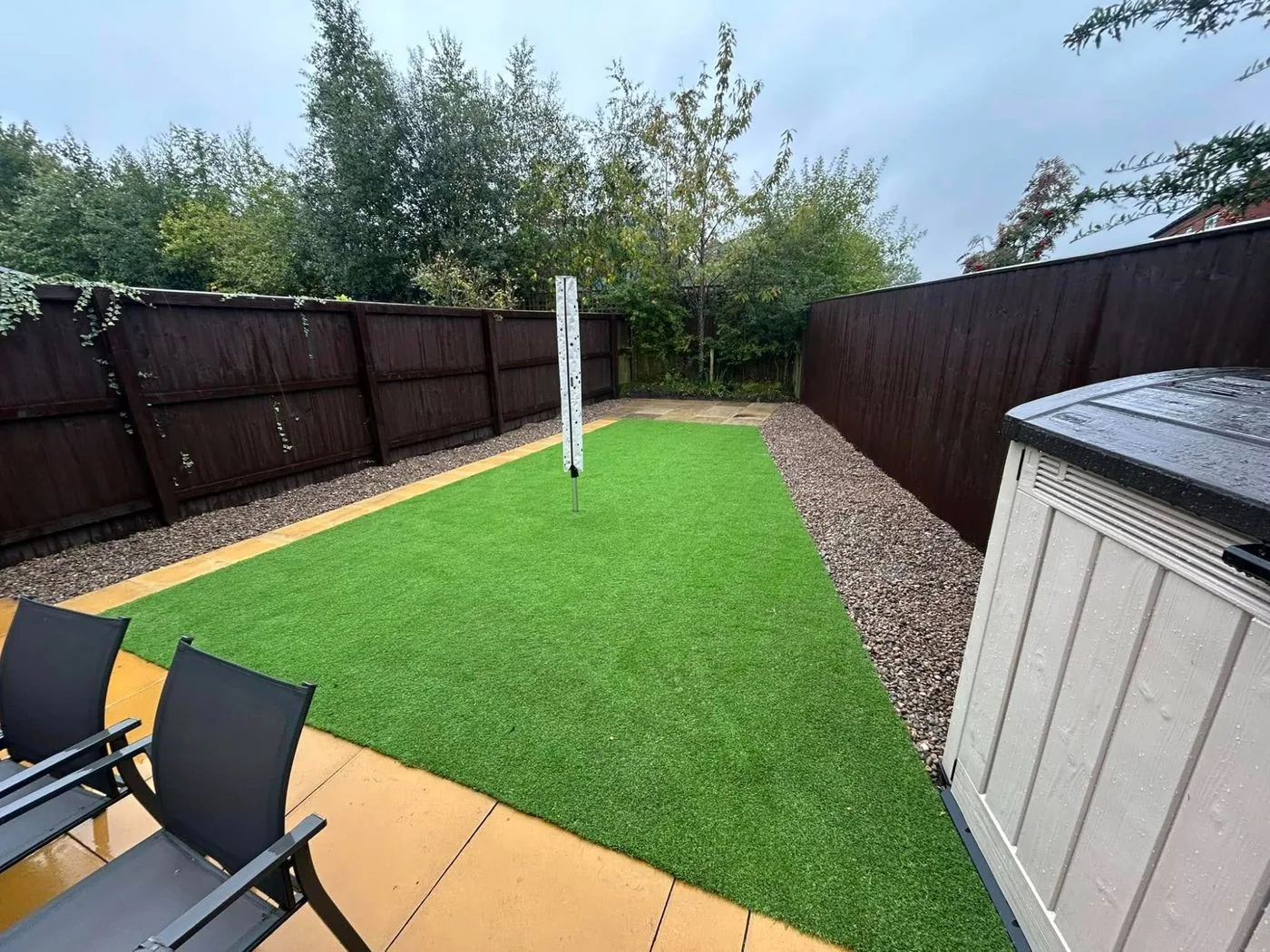 Rectangular artificial lawn with tan paving, gravel borders and dark garden fencing